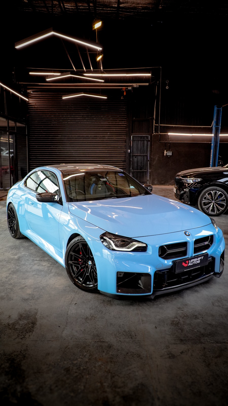 A light blue BMW M2 with black wheels, front three-quarter view in the workshop at night.