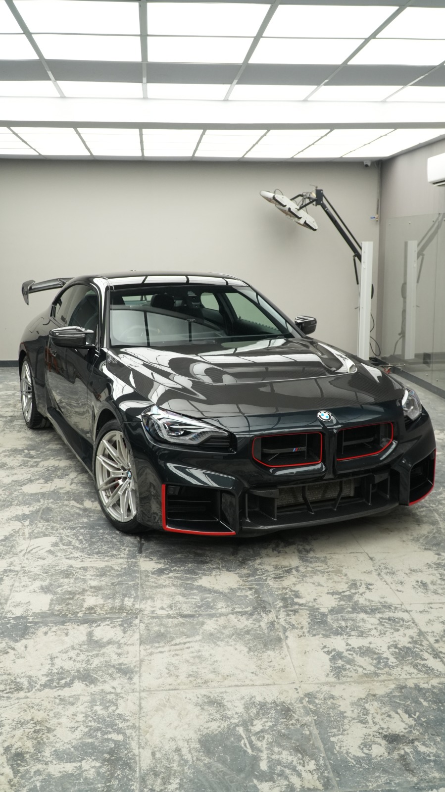 The black BMW M2 with red accents on its grille and splitter, front three-quarter view in a white-walled bay.