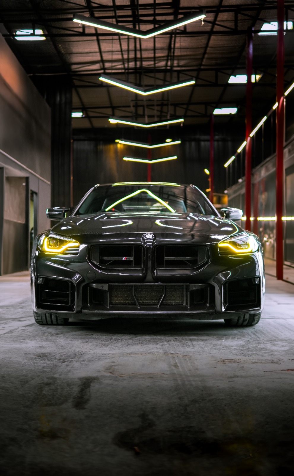 The black BMW M2 head-on in the workshop hall, yellow DRLs lit beneath the chevron strip lights.