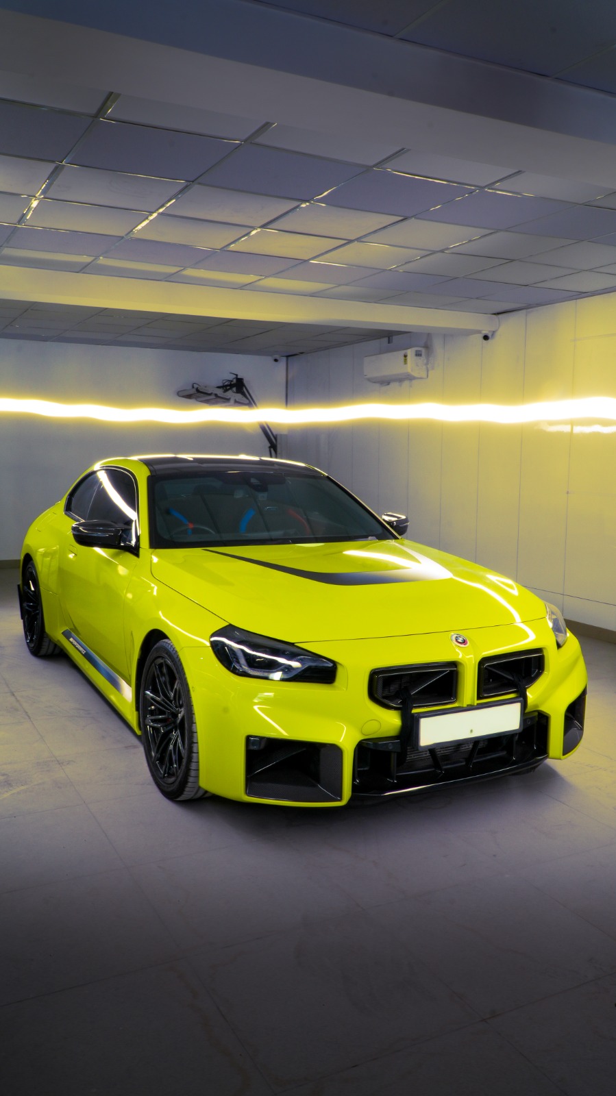 A bright yellow BMW M2 with black wheels, front three-quarter view in a bay lit by a yellow light strip.