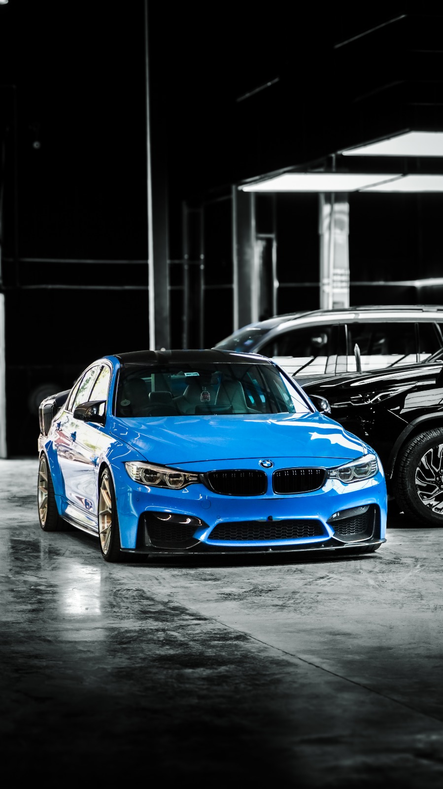 A blue BMW M3 saloon with bronze wheels, front three-quarter view on a wet workshop floor.