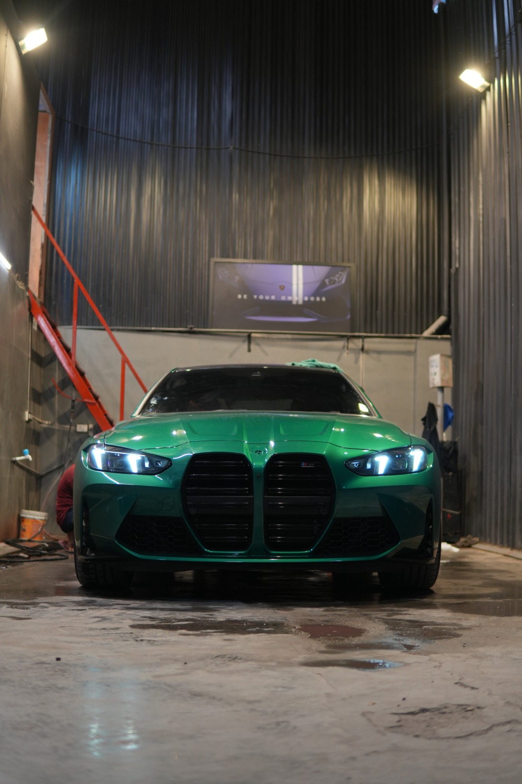 The satin green BMW M4 head-on inside a corrugated-walled bay, its tall kidney grille and headlamps lit.