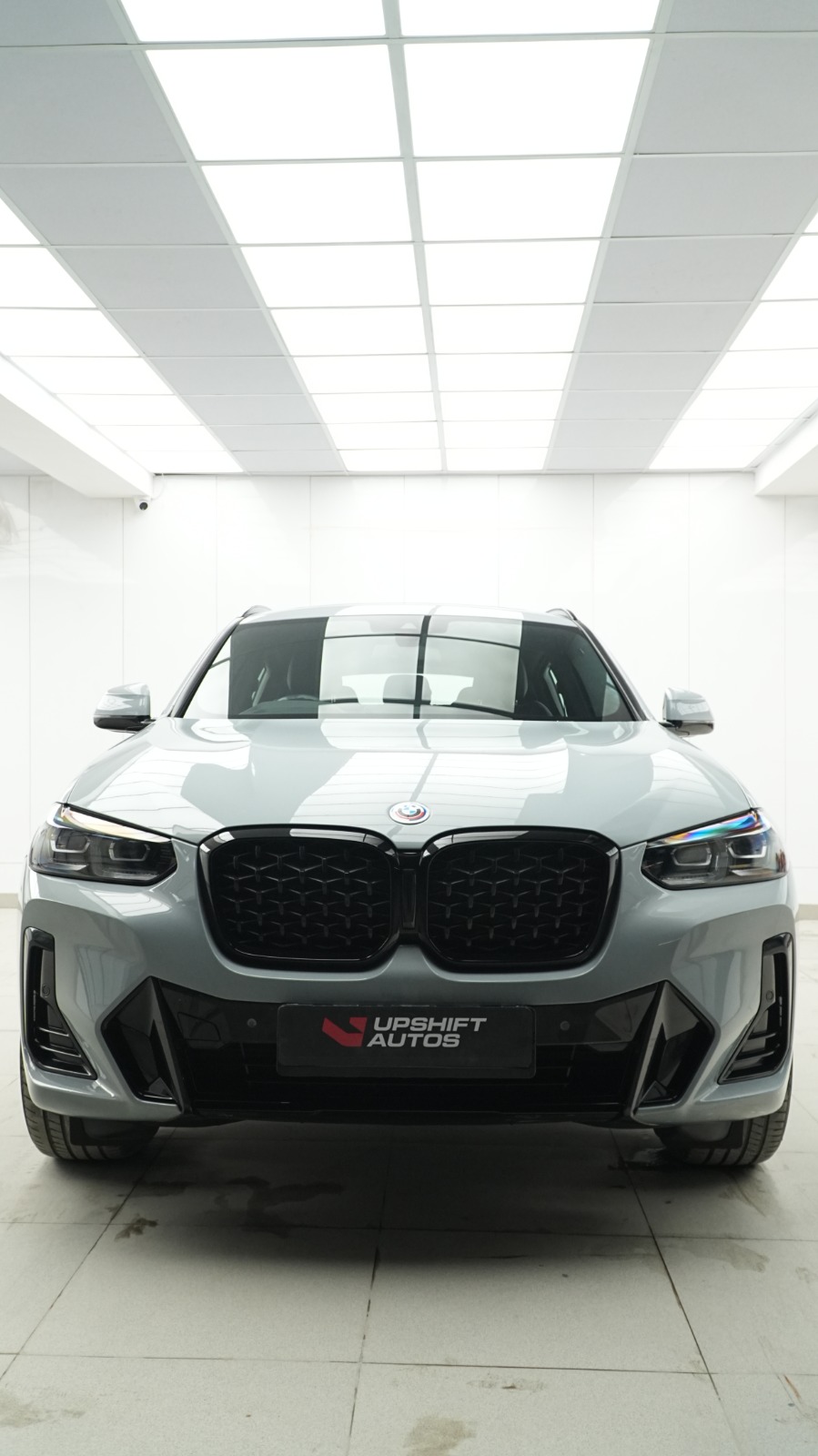 A grey BMW X3 head-on in a bright white bay with ceiling light panels.