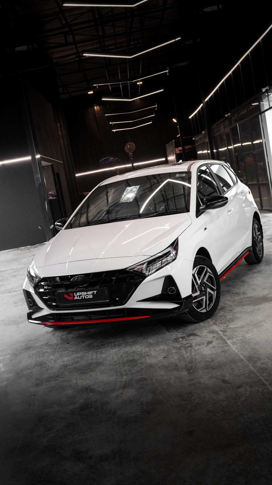 A white Hyundai i20 N Line with red trim along its lip, front three-quarter view in the workshop hall.