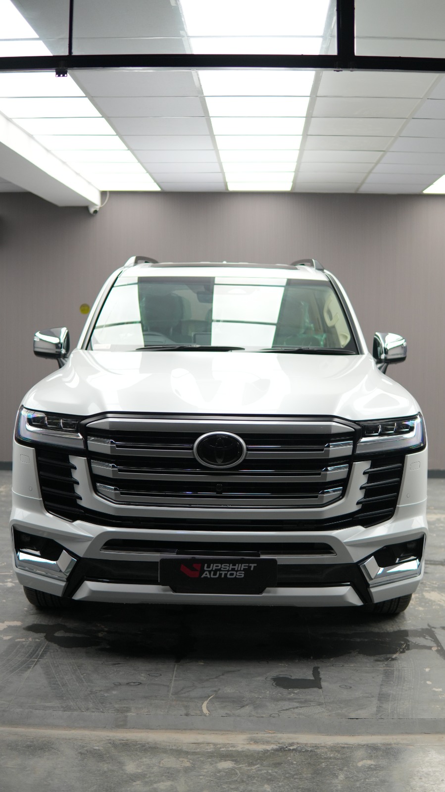 A white Toyota Land Cruiser LC300 with a chrome grille, head-on in a bright white bay.