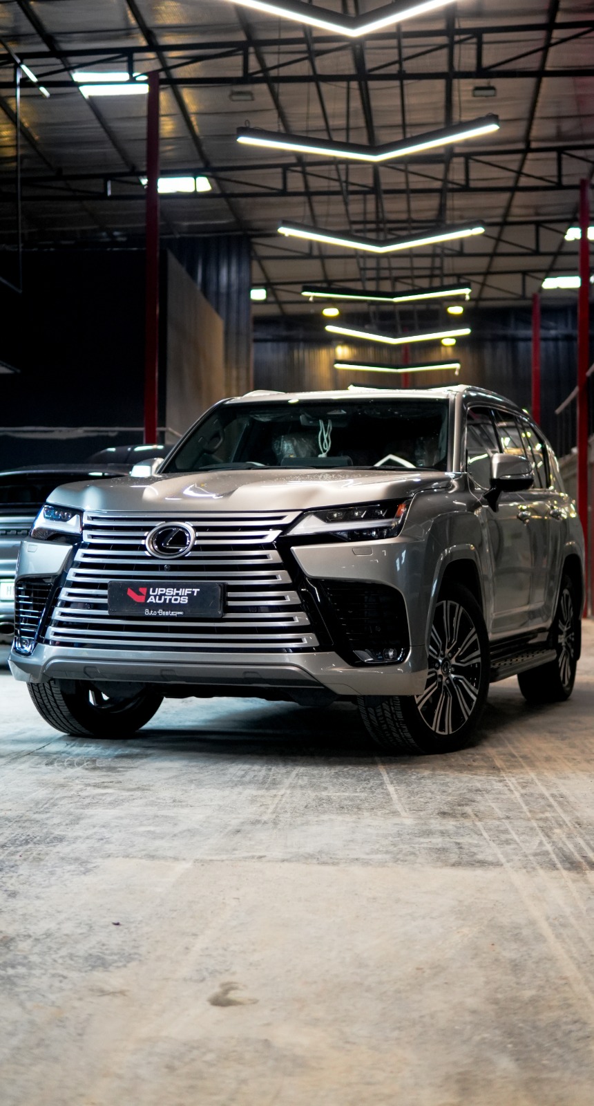 The silver Lexus LX head-on in the workshop hall under strip lights, its chrome grille bars catching the light.