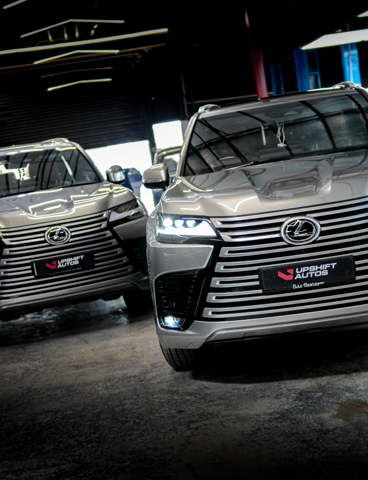 Two silver Lexus LX SUVs side by side in the workshop, the nearer one close to the camera with its headlamps lit.