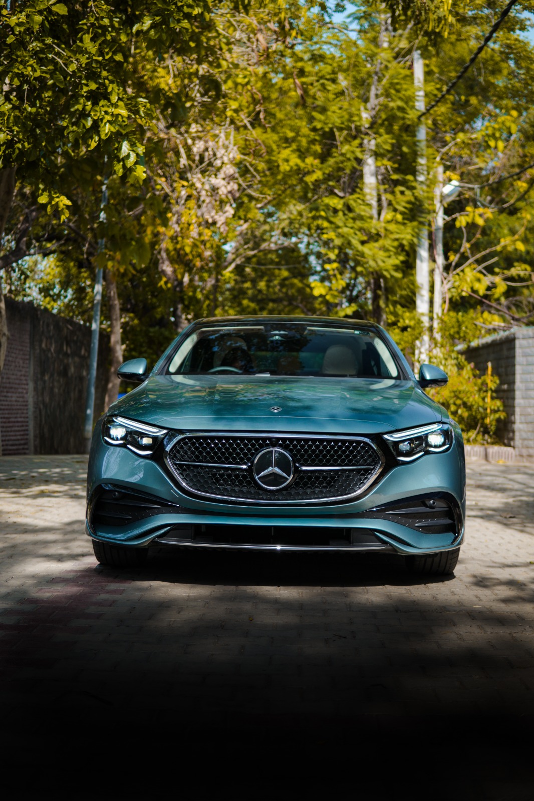 The grey-green Mercedes-Benz E-Class head-on on a lane lined with trees, headlamps lit.