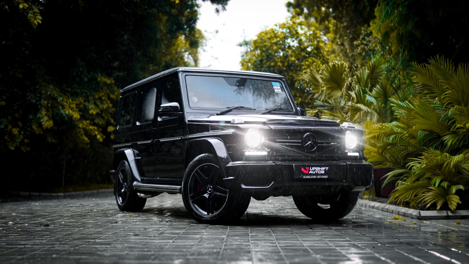 The black G63 outdoors on a paved drive among palms, headlamps lit.