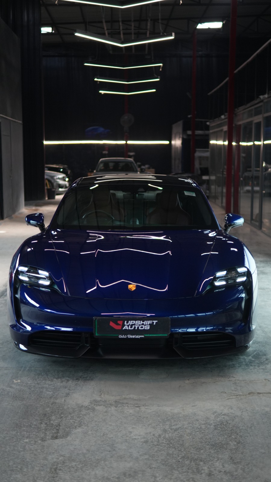 A deep blue Porsche Taycan head-on in the workshop hall, headlamps lit beneath angled strip lights.
