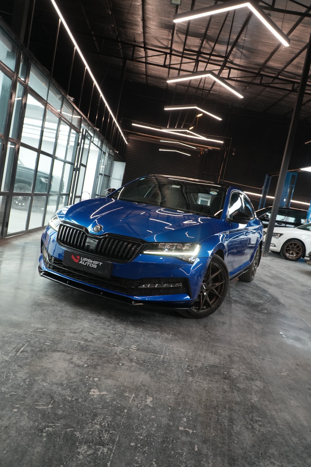 A blue Škoda Superb with black wheels, front three-quarter view in the workshop hall.