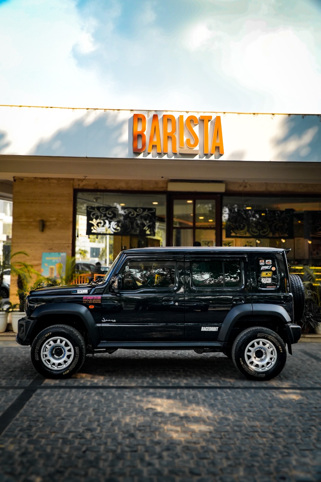 A black five-door Maruti Suzuki Jimny with silver steel wheels, side view outside a café.