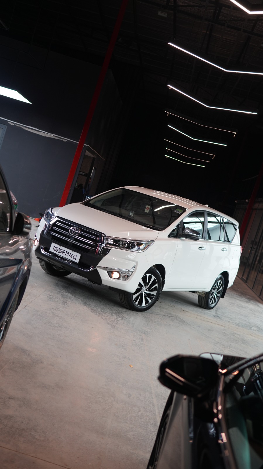The white Toyota Innova Crysta from a low angle in the workshop hall, beside the door mirror of another car.